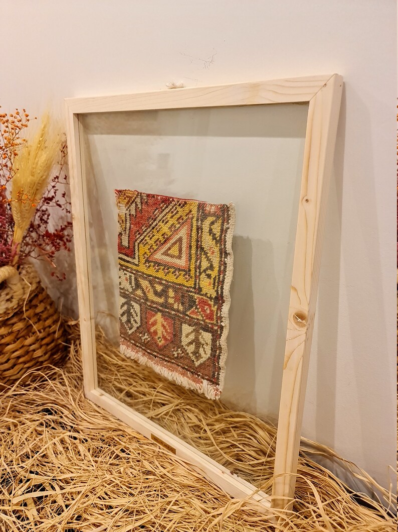 May include: A small, colourful rug with geometric patterns is framed in a natural wood frame with a clear glass front.