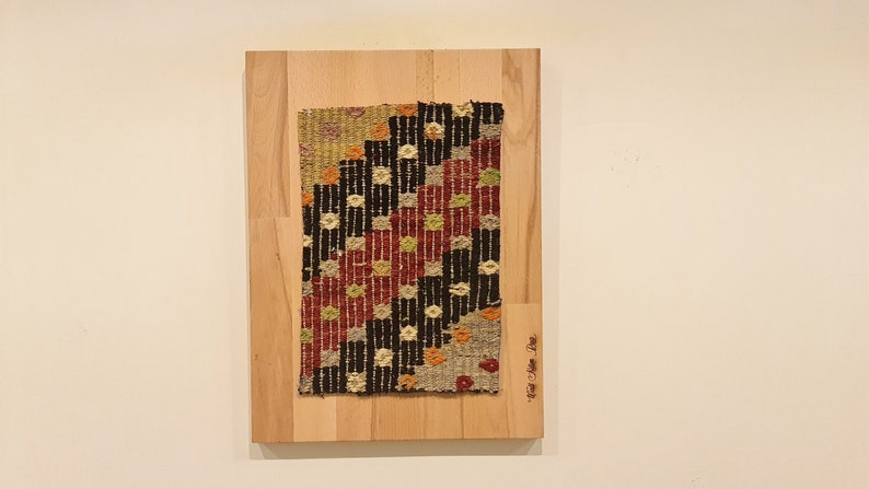 Puede incluir: Un tapiz tejido enmarcado con un patr&oacute;n geom&eacute;trico en tonos rojos, negros, blancos y verdes. El tapiz est&aacute; montado en una tabla de madera con el texto "The Art of the Loom" escrito en la esquina inferior derecha.