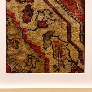 May include: Close-up of a framed rug with a detailed pattern of red, brown, and beige. The rug has a geometric design with intricate details.