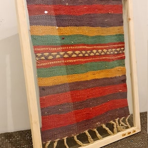 May include: A framed woven tapestry with colorful stripes in shades of red, green, yellow, and purple. The tapestry has a fringe border and is framed in a natural wood frame.