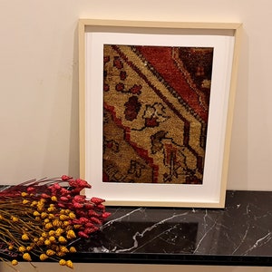 May include: A framed print of a vintage rug with a red, brown, and beige pattern. The frame is a light wood color and the print is on a white background. The print is sitting on a black marble table.