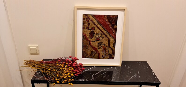 May include: A framed print of a vintage rug with a red, brown, and beige pattern. The frame is a light wood color and the print is on a white background. The print is sitting on a black marble table.