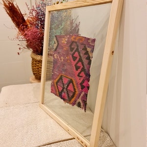 May include: A framed piece of colorful woven fabric with a geometric pattern in shades of pink, purple, orange, and black. The frame is made of natural wood and has a glass front.