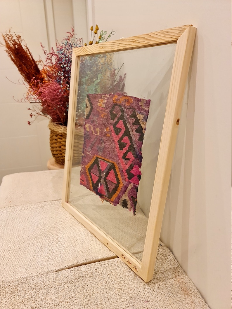 May include: A framed piece of colorful woven fabric with a geometric pattern in shades of pink, purple, orange, and black. The frame is made of natural wood and has a glass front.