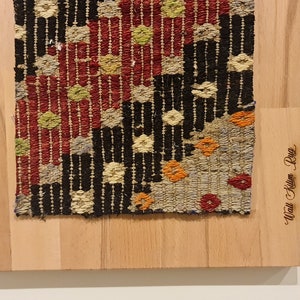 Puede incluir: Un primer plano de una alfombra tejida colorida con un patr&oacute;n geom&eacute;trico. La alfombra est&aacute; enmarcada en madera y tiene el texto "Afghan Kilim Rug" grabado en el marco.