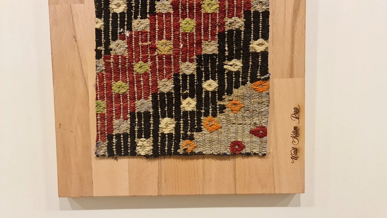 Puede incluir: Un primer plano de una alfombra tejida colorida con un patr&oacute;n geom&eacute;trico. La alfombra est&aacute; enmarcada en madera y tiene el texto "Afghan Kilim Rug" grabado en el marco.