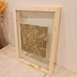 May include: A framed piece of woven fabric with a geometric pattern in shades of brown, beige, and pink. The fabric is displayed in a simple wooden frame with a glass front.