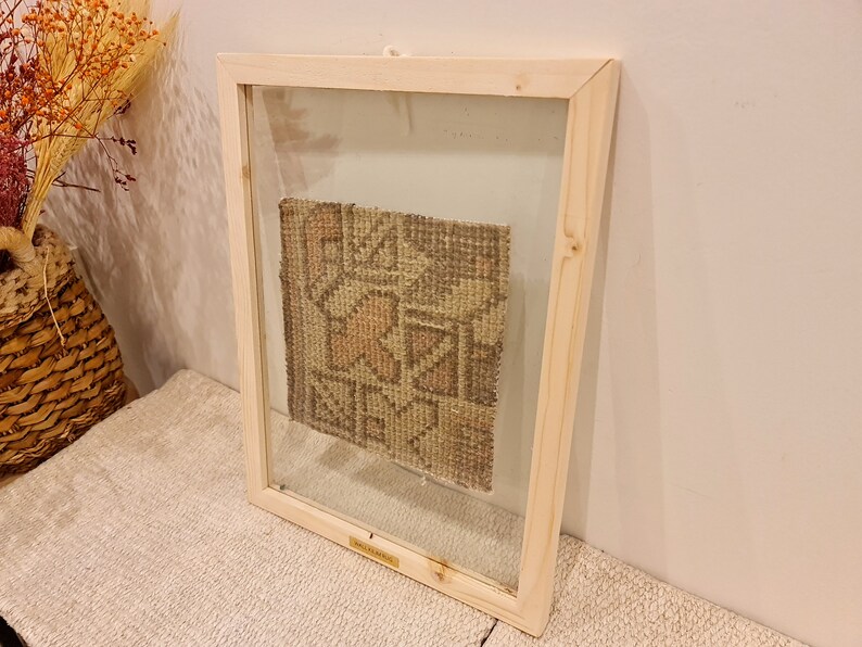 May include: A framed piece of woven fabric with a geometric pattern in shades of brown, beige, and pink. The fabric is displayed in a simple wooden frame with a glass front.