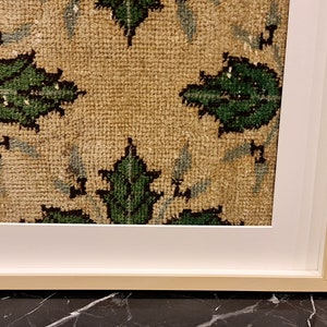 May include: A framed piece of art featuring a beige fabric with a repeating pattern of green leaves. The fabric is framed in a white mount and a natural wood frame.