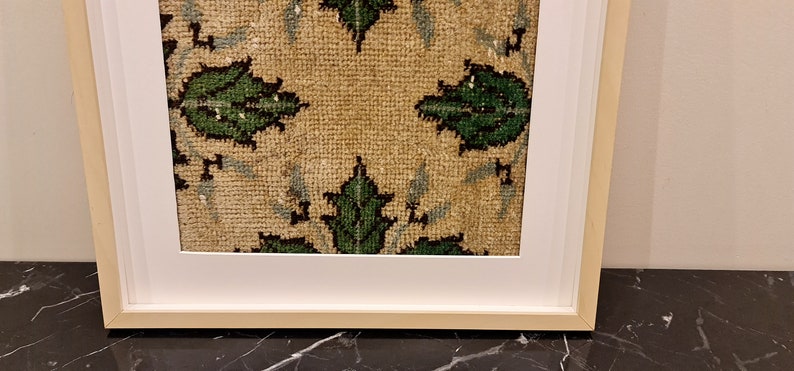 May include: A framed piece of art featuring a beige fabric with a repeating pattern of green leaves. The fabric is framed in a white mount and a natural wood frame.