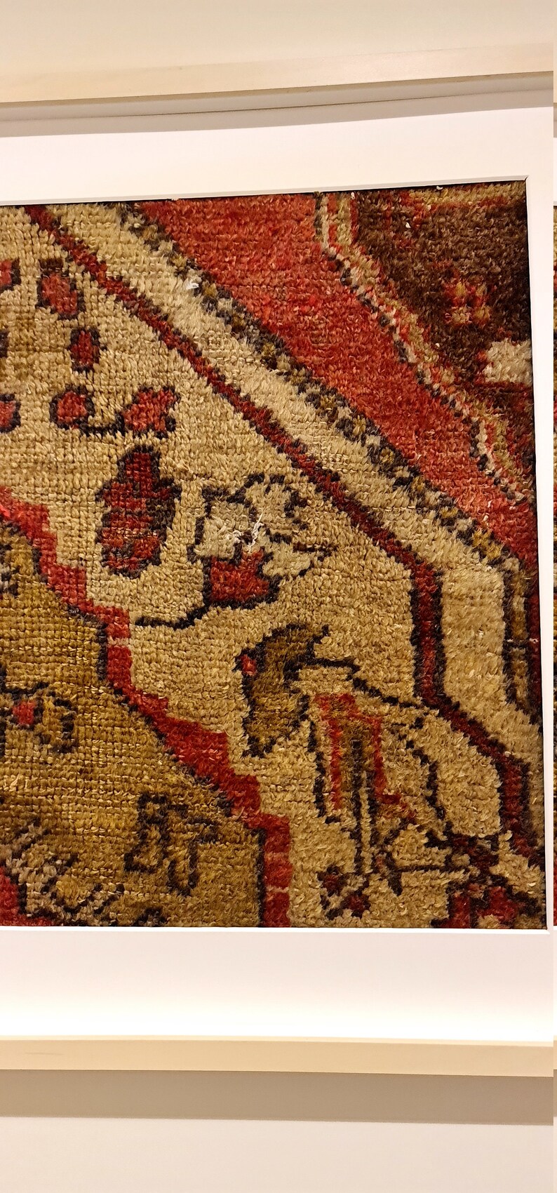 May include: Close-up of a red, brown, and beige patterned rug with intricate designs. The rug has a textured surface and is framed in a white frame.