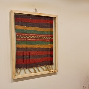 May include: A framed multi-colored woven tapestry with a fringe. The tapestry is made of different shades of red, orange, yellow, green, and brown.