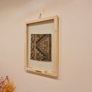May include: A framed piece of woven fabric with a geometric pattern in shades of brown, yellow, and pink. The fabric is displayed in a simple wooden frame with a glass front.