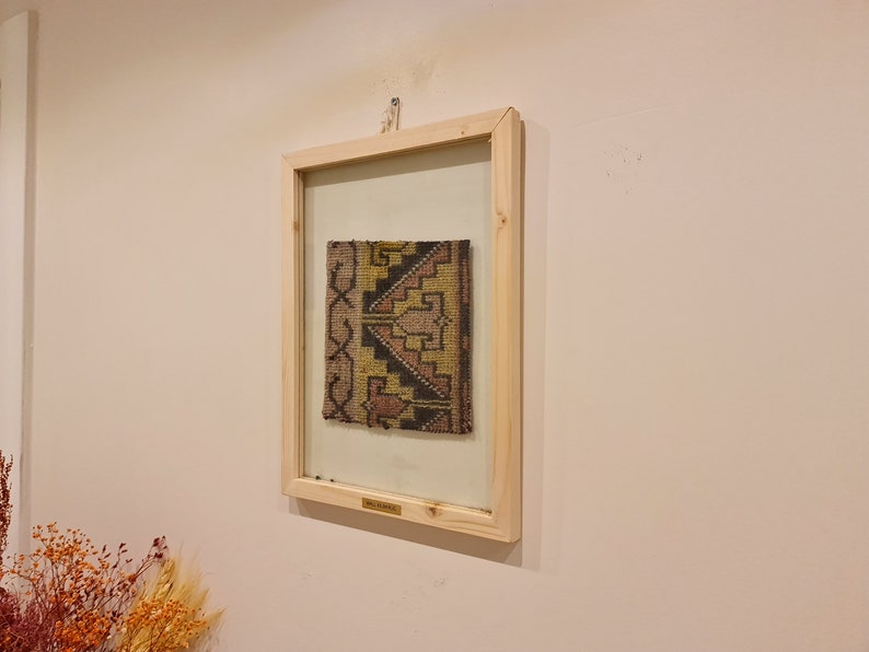 May include: A framed piece of woven fabric with a geometric pattern in shades of brown, yellow, and pink. The fabric is displayed in a simple wooden frame with a glass front.