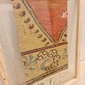 May include: A framed vintage rug with a cream background and a pink, brown, and blue floral pattern. The rug has fringe on the bottom and is labelled "Wall Rug".