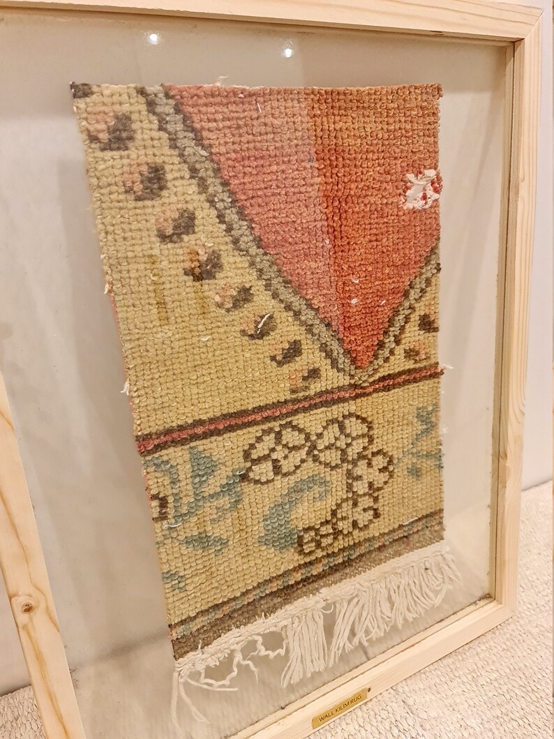 May include: A framed vintage rug with a cream background and a pink, brown, and blue floral pattern. The rug has fringe on the bottom and is labelled "Wall Rug".