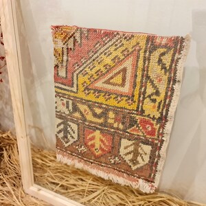 May include: A small, framed rug with a colourful geometric pattern in shades of red, yellow, brown, and white. The rug has a fringe border and is displayed in a wooden frame.