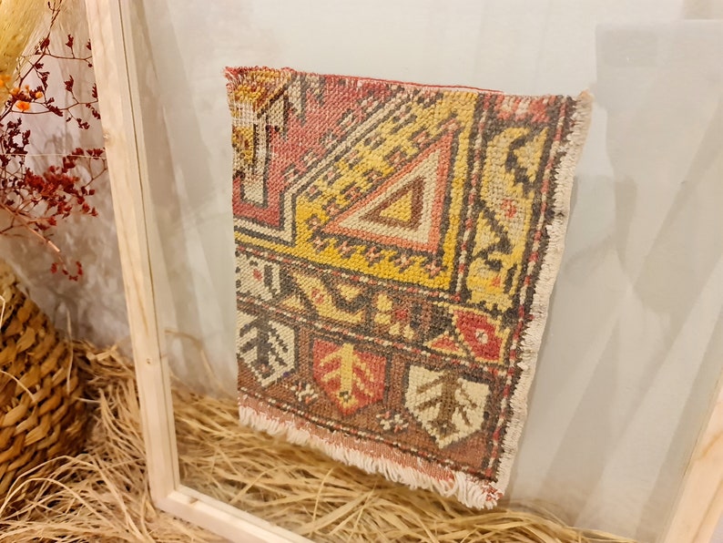 May include: A small, framed rug with a colourful geometric pattern in shades of red, yellow, brown, and white. The rug has a fringe border and is displayed in a wooden frame.