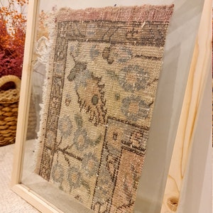 May include: A framed vintage rug with a faded floral pattern in shades of pink, blue, and beige. The rug is displayed in a wooden frame with a glass front. The frame has a gold label that reads "WALL RUG".