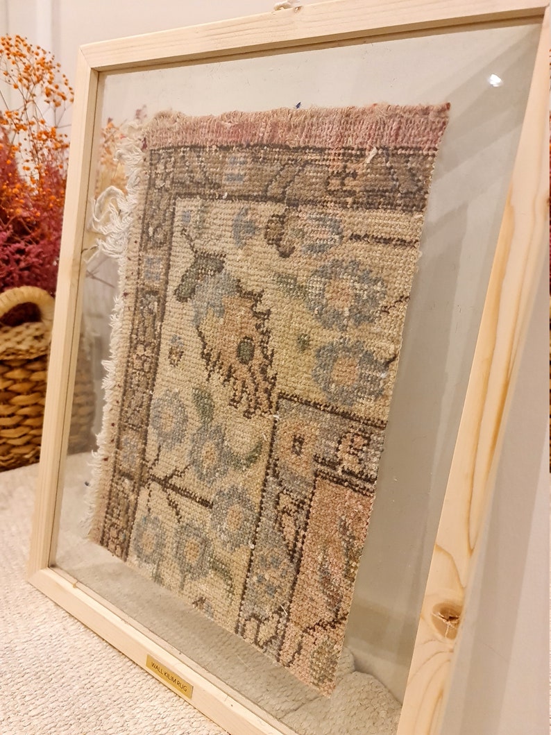 May include: A framed vintage rug with a faded floral pattern in shades of pink, blue, and beige. The rug is displayed in a wooden frame with a glass front. The frame has a gold label that reads "WALL RUG".