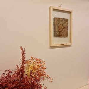 May include: A framed piece of woven fabric with a geometric pattern in shades of brown, beige, and orange. The fabric is displayed against a white wall.