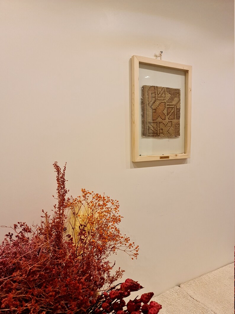 May include: A framed piece of woven fabric with a geometric pattern in shades of brown, beige, and orange. The fabric is displayed against a white wall.
