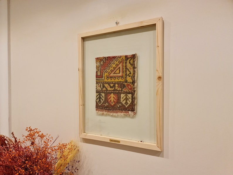 May include: A framed piece of antique rug with a geometric pattern in shades of red, yellow, brown, and white. The rug is framed in a natural wood frame.