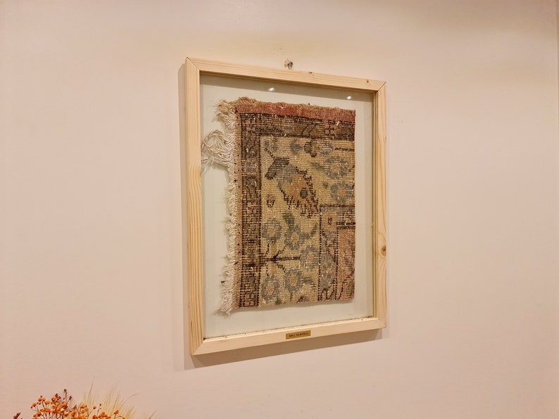 May include: A framed piece of antique rug with a faded floral pattern in shades of beige, brown, and pink. The rug is framed in a natural wood frame with a glass front.
