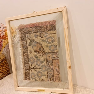 May include: A framed piece of vintage rug with a faded floral pattern in shades of pink, blue, and brown. The rug is framed in a natural wood frame with a glass front.