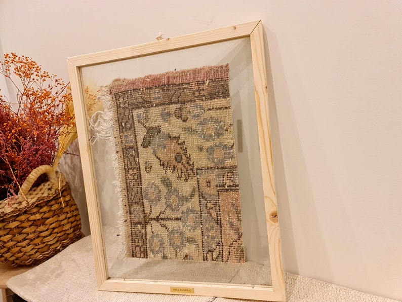 May include: A framed piece of vintage rug with a faded floral pattern in shades of pink, blue, and brown. The rug is framed in a natural wood frame with a glass front.