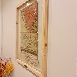 May include: A framed piece of colourful woven fabric with fringe hanging from the bottom. The fabric is a mix of red, yellow, and blue colours. The frame is made of natural wood.