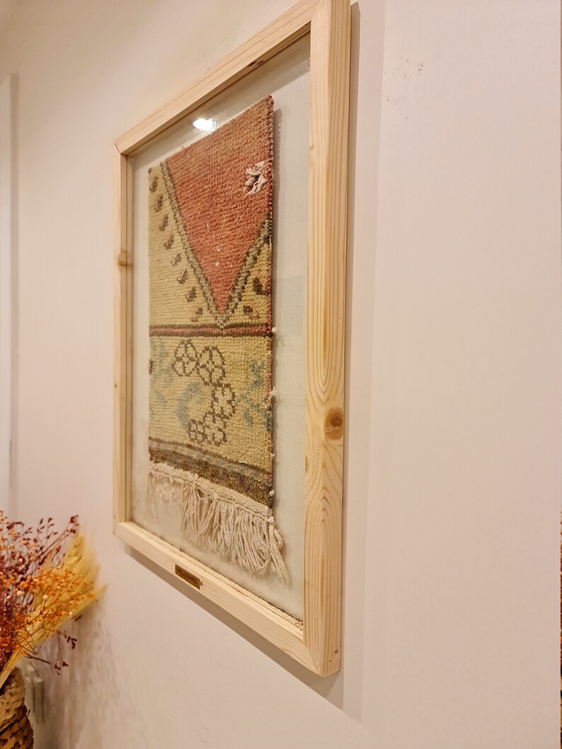 May include: A framed piece of colourful woven fabric with fringe hanging from the bottom. The fabric is a mix of red, yellow, and blue colours. The frame is made of natural wood.