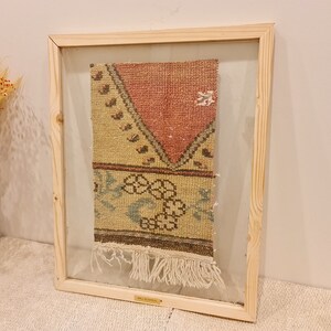 May include: A framed vintage rug with a geometric pattern in shades of pink, beige, and blue. The rug has a fringe border and is displayed in a wooden frame.