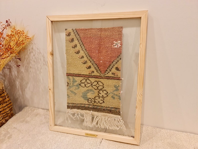 May include: A framed vintage rug with a geometric pattern in shades of pink, beige, and blue. The rug has a fringe border and is displayed in a wooden frame.