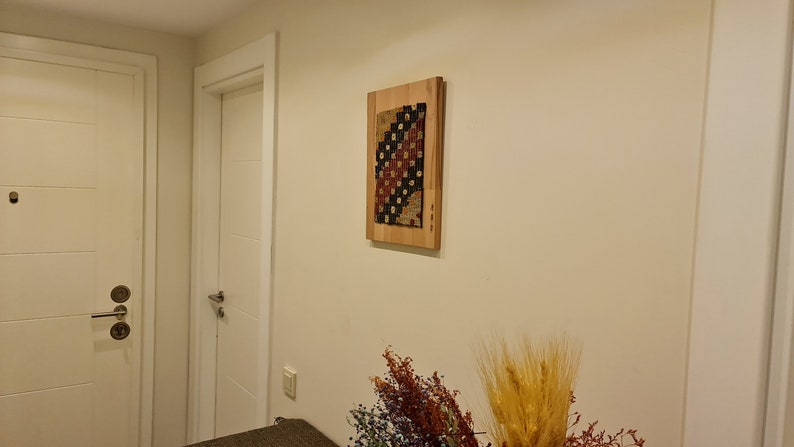 Puede incluir: Un tapiz enmarcado con un patr&oacute;n geom&eacute;trico en tonos rojos, negros y blancos, colgado en una pared blanca en un pasillo.