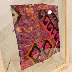 May include: A framed piece of colorful woven fabric with geometric patterns in shades of pink, purple, orange, and black. The fabric is displayed in a wooden frame with a glass front.