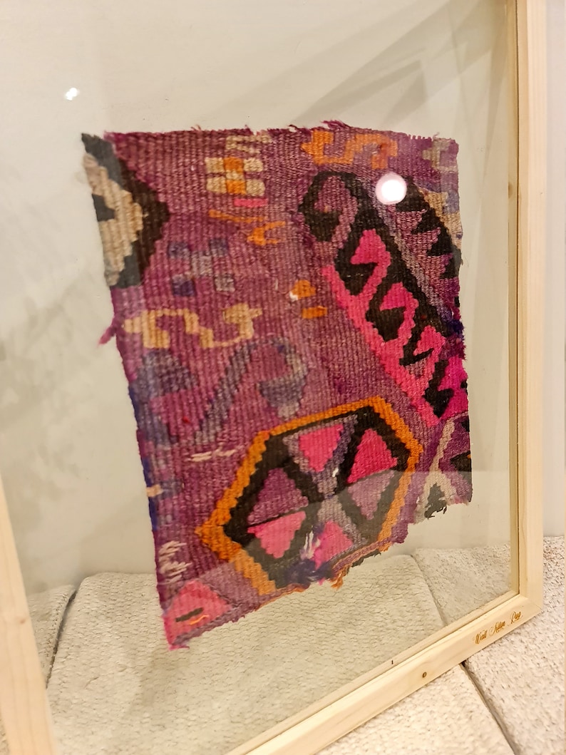 May include: A framed piece of colorful woven fabric with geometric patterns in shades of pink, purple, orange, and black. The fabric is displayed in a wooden frame with a glass front.