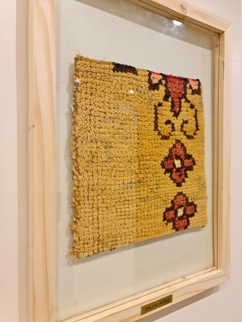 Puede incluir: Una alfombra kilim amarilla y roja enmarcada con un patr&oacute;n floral. El marco es de madera natural y tiene una etiqueta que dice "Alfombra kilim de pared".