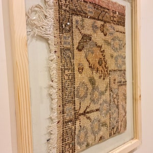 May include: A framed vintage rug with a faded floral pattern in shades of brown, beige, and blue. The rug has a frayed edge and is displayed in a simple wooden frame.