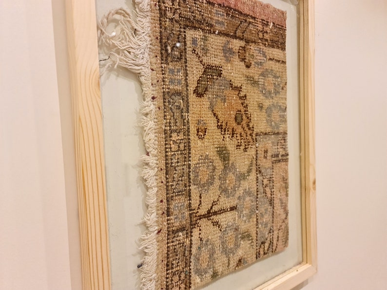 May include: A framed vintage rug with a faded floral pattern in shades of brown, beige, and blue. The rug has a frayed edge and is displayed in a simple wooden frame.