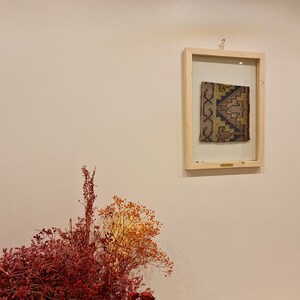 May include: A framed piece of fabric with a geometric pattern in shades of brown, yellow, and black. The fabric is displayed against a white wall with dried flowers in the foreground.