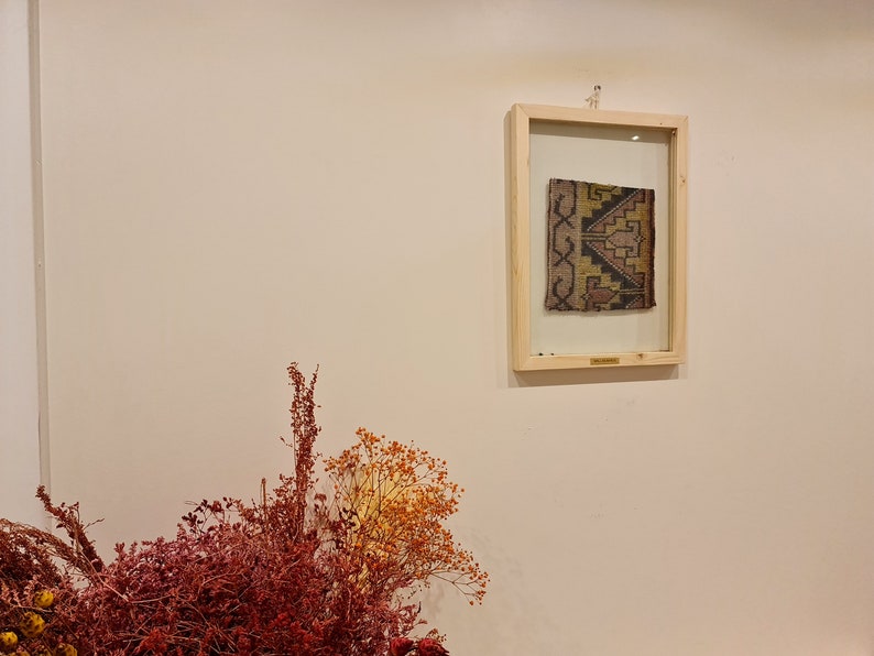 May include: A framed piece of fabric with a geometric pattern in shades of brown, yellow, and black. The fabric is displayed against a white wall with dried flowers in the foreground.
