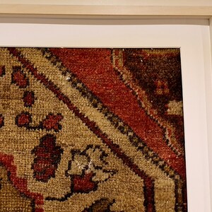 May include: A framed piece of art featuring a close-up of a colorful rug with intricate patterns. The rug is primarily beige with red, brown, and black accents.