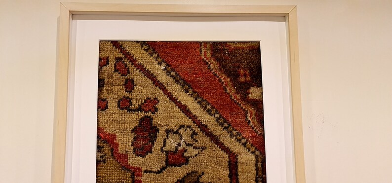 May include: A framed piece of art featuring a close-up of a colorful rug with intricate patterns. The rug is primarily beige with red, brown, and black accents.