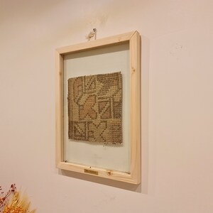 May include: A framed piece of woven fabric with a geometric pattern in shades of brown and beige. The frame is made of natural wood and has a glass front.