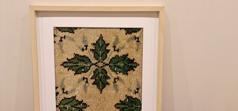 May include: A framed piece of art featuring a beige background with a repeating pattern of green leaves. The frame is a light wood colour with a white mount.