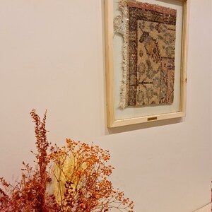 May include: A framed piece of antique rug with a floral pattern in shades of brown, pink, and blue. The rug is displayed on a white wall with dried flowers in the foreground.