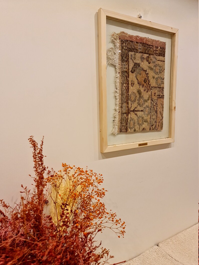 May include: A framed piece of antique rug with a floral pattern in shades of brown, pink, and blue. The rug is displayed on a white wall with dried flowers in the foreground.