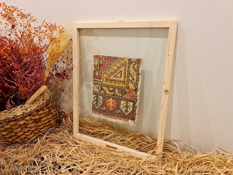 May include: A framed piece of colourful patterned fabric with a fringe border, displayed against a light brown background.