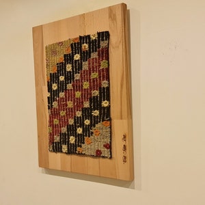 Puede incluir: Una alfombra tejida colorida con patrones geom&eacute;tricos en tonos rojos, negros, blancos y amarillos est&aacute; montada en una tabla de madera. La tabla tiene el texto "Hand Woven Rug" inscrito en ella.